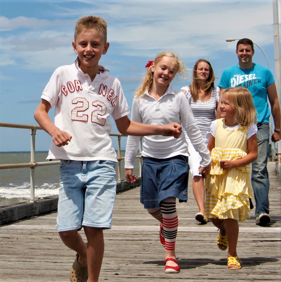 Two parents in background with three children smiling and walking in front of them on a pier